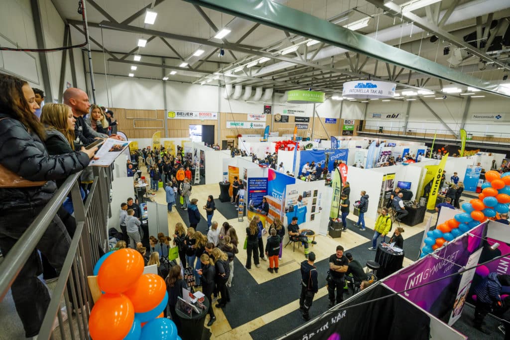 Mässa i en stor hall med mässmontrar, besökare och utställare sett uppifrån, samt en balkong där det står några personer och tittar ner.