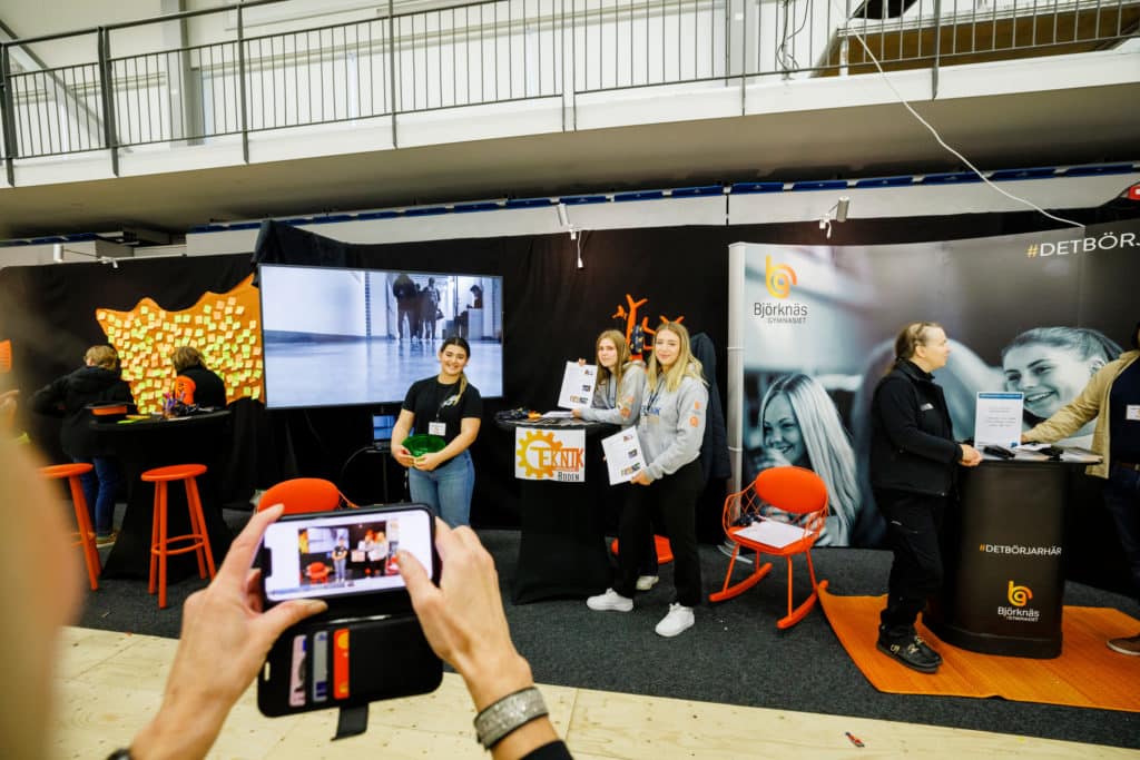 Exhibition stand in black and orange, a mobile camera in front.
