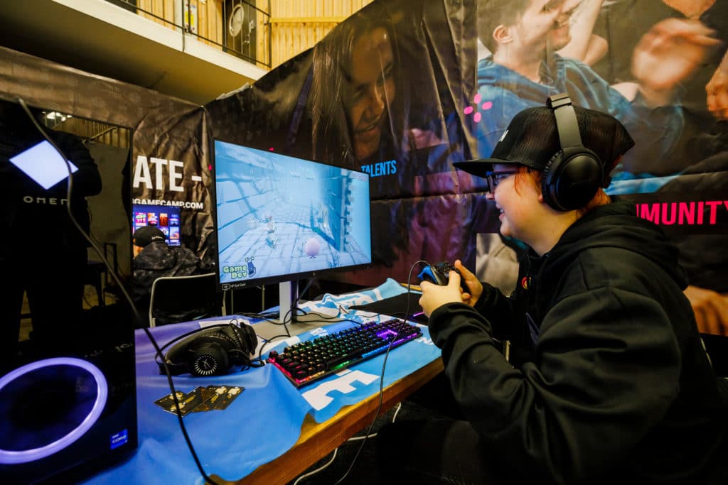Young person in cap and headphones holds a controller in front of a computer screen showing a game.