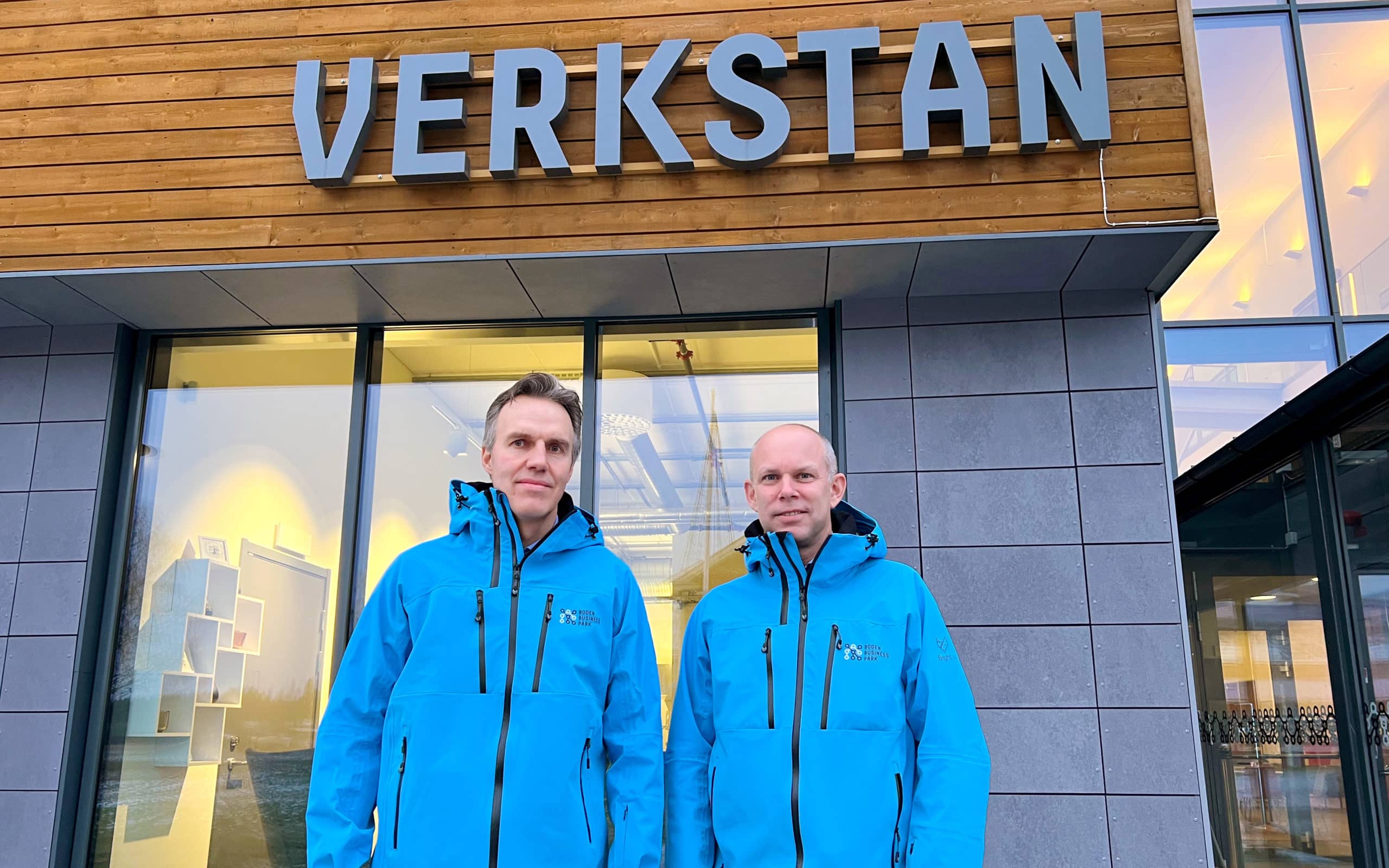 Bild på Håkan Nordin och Nils Lindh som har. på sig en blå jacka och står framför en byggnad med en skylt där det står Verkstan. Båda ser glada ut och tittar in i kameran. 