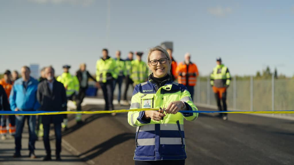 A woman in a reflective jacket cuts a ribbon at an inauguration. In the background is a group of people in different colorful work clothes. The sky is clear blue.