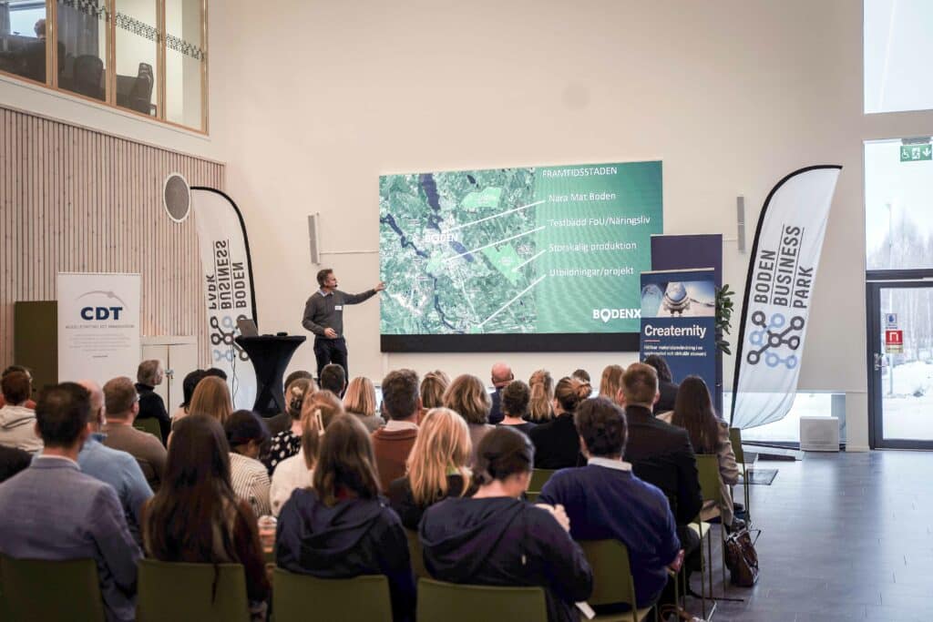 A speaker presents in front of a large screen with a satellite image and the text "City of the Future" at an event. The audience, sitting in a lecture hall, listens attentively. The event seems to be related to local initiatives in Boden, such as local food and large-scale production. Banners for Boden Business Park and CDT, together with the Bodenxt and Creaternity logos, suggest a focus on innovation and business development.
