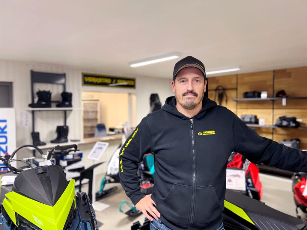 A man is standing inside a showroom with snowmobiles and related equipment. He is wearing a black jacket with yellow accents and a cap with a logo, probably representing the brand of the snowmobiles. The interior is well-lit and suggests a commercial or retail environment.