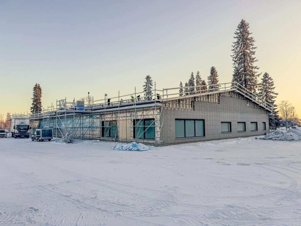 A winter landscape of a construction site with a single-storey building partly covered by scaffolding. Snow covers the ground, and some conifers stand in the background, bathed in the soft light of a setting or rising sun.