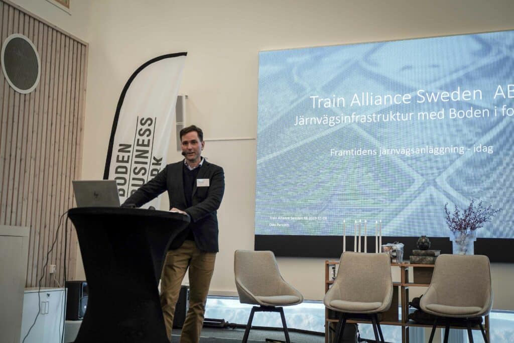 Figure 2: A man stands at a lectern with a presentation behind him showing "Train Alliance Sweden AB" and the title "Railway infrastructure with Boden in focus".
