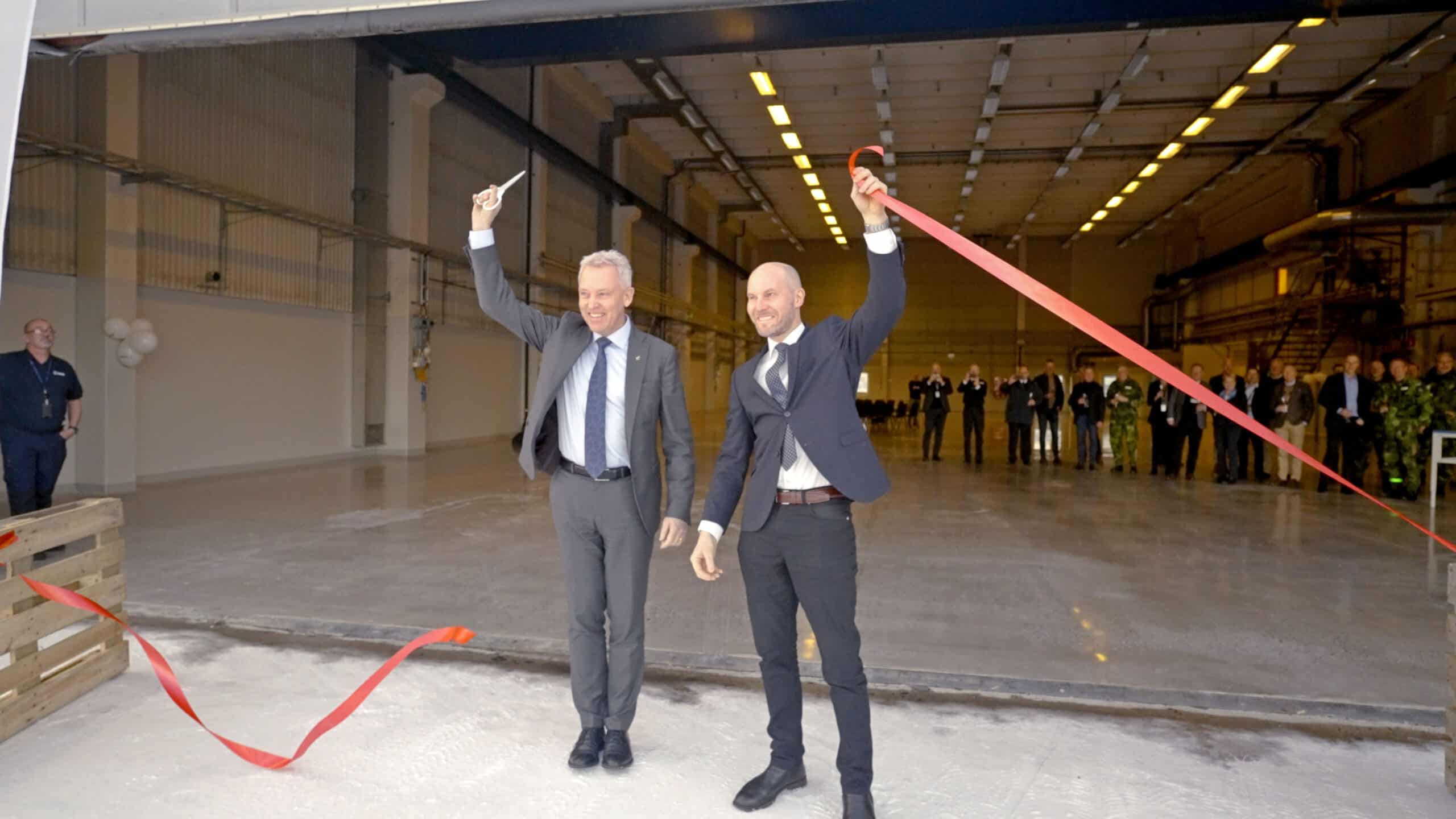 Två leende män i kostym klipper ett rött band vid en invigningsceremoni inne i en stor industrihall med åskådare i bakgrunden.