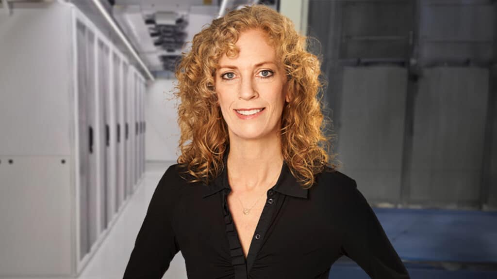 The image shows a woman with curly hair standing in front of server racks in a data center. She is wearing a black shirt and appears to be in a professional role, possibly in IT or managing a technology department. The background shows a clean and well-organized server environment, indicating a modern and high-tech workplace.