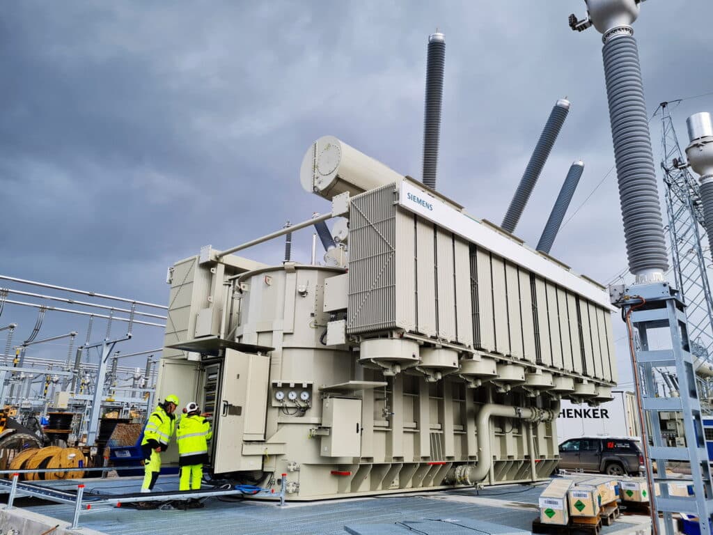 Tekniker i säkerhetskläder inspekterar en stor transformator märkt Siemens på ett elnätställverk under en molnig himmel.