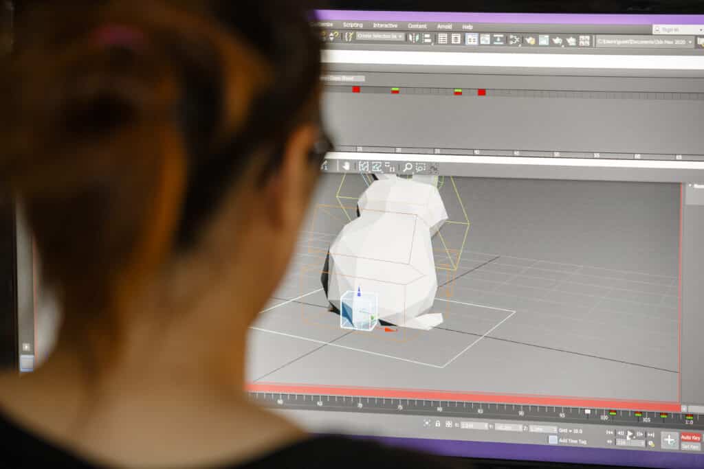 A person looking at a computer screen showing a 3D model in a design program. The person is seen from behind with the focus on the screen showing a polygon model in a software environment.