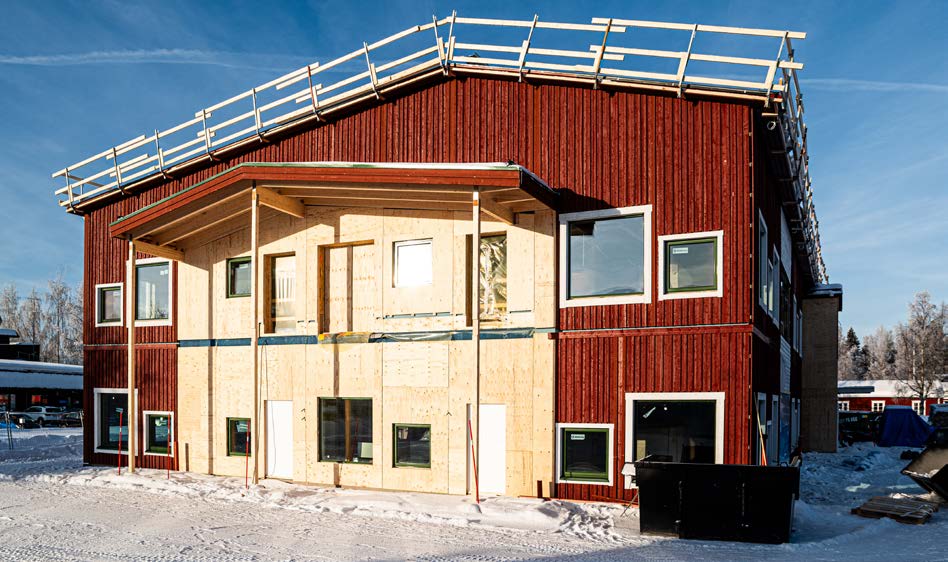 En tvåvåningsbyggnad under konstruktion med rödmålad träfasad och exponerad träpanel på framsidan. Byggnaden är omgiven av snö och byggställningar syns på taket under en klar blå himmel.