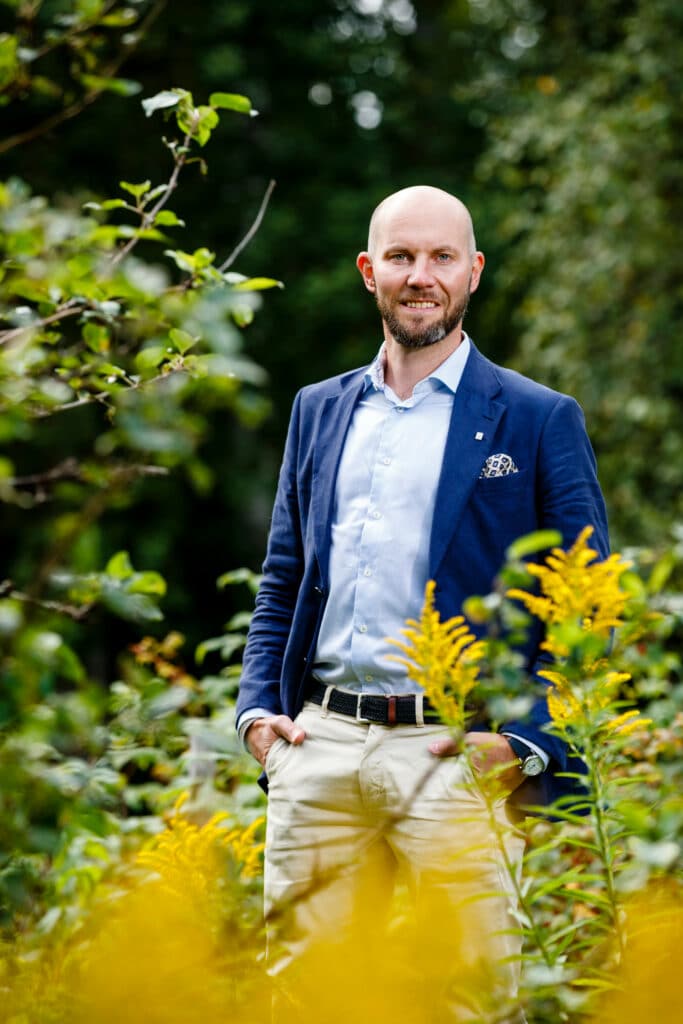 En man i blå kavaj och ljusa byxor står utomhus i en grönskande miljö, omgiven av växter och gula blommor. Han är flintskallig, har skägg och ler medan han står med händerna i fickorna. Bakgrunden är fylld av grönska, vilket ger bilden en naturlig och fridfull känsla.