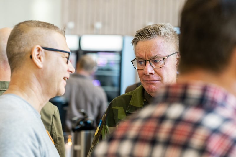 Två män pratar under en paus vid ett företagsevent. En av männen är iklädd en militäruniform med glasögon, medan den andra mannen är i en mer avslappnad klädstil. Bakgrunden är något suddig med människor som minglar.