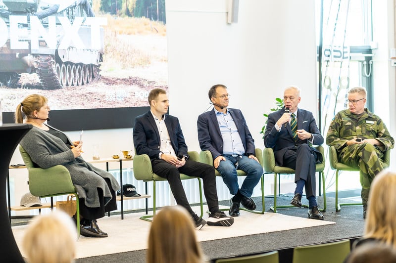 A panel discussion is taking place during a corporate event. Five people are seated in a row on a stage, one wearing a military uniform and the others dressed in civilian clothes. A background image shows a tank and the text "Bodenxt." 