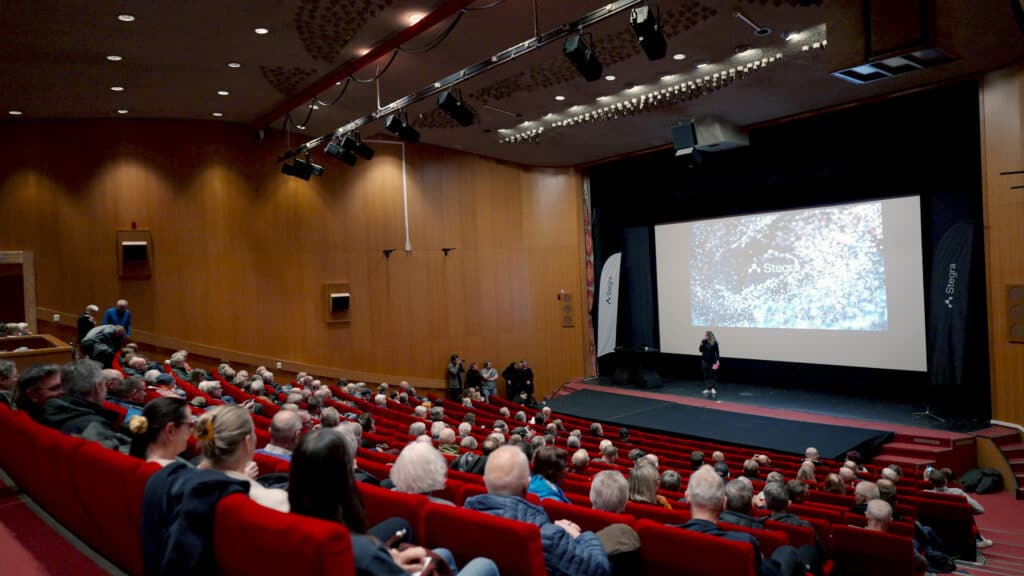 Halvfylld salong på Folkets Hus i Boden under en frågestund med Stegra. På scen står Henrik Henriksson, vd för Stegra, framför en stor skärm med företagets logotyp.