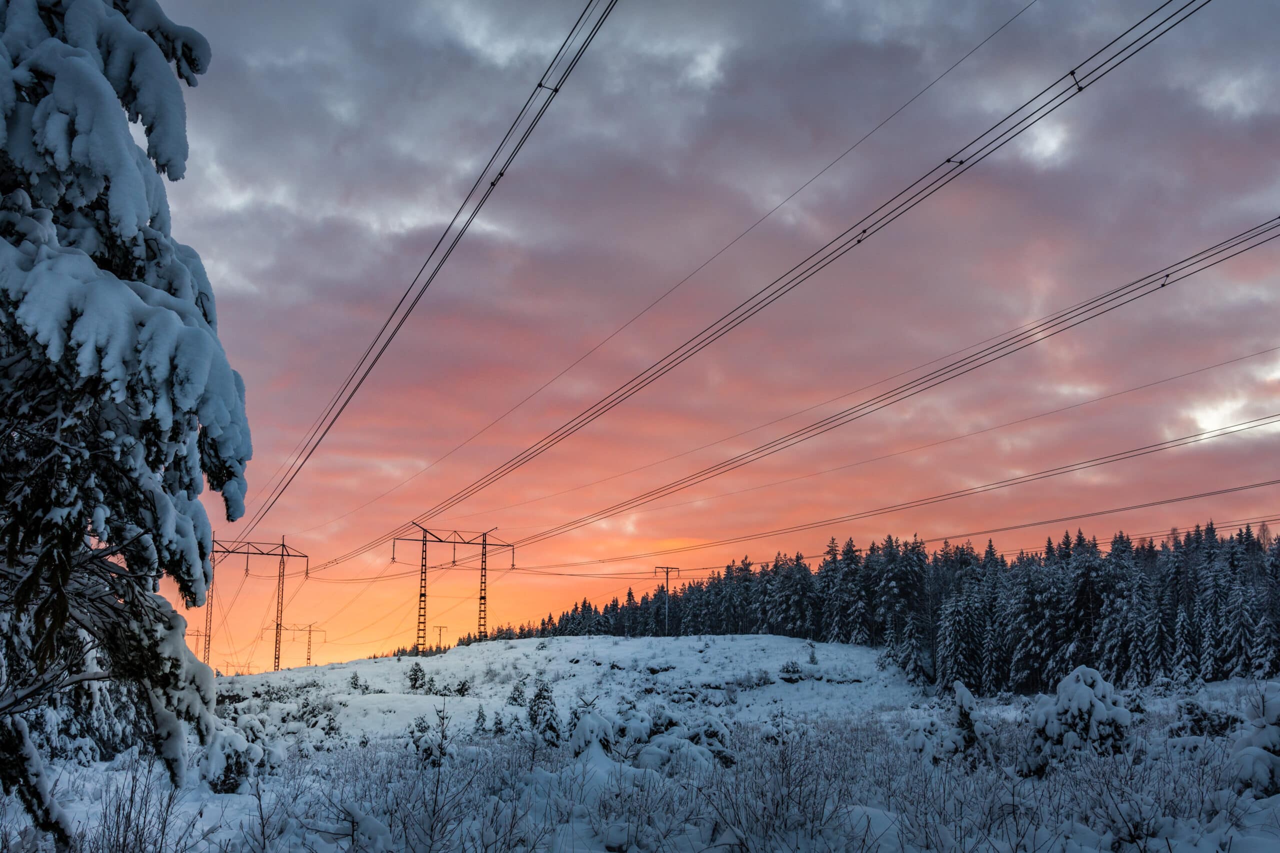 Högspänningsledningar som sträcker sig över ett snötäckt vinterlandskap, med en solnedgång i bakgrunden som färgar himlen i rosa och orange toner. Snötäckta träd omger området, vilket skapar en naturskön och fridfull atmosfär.