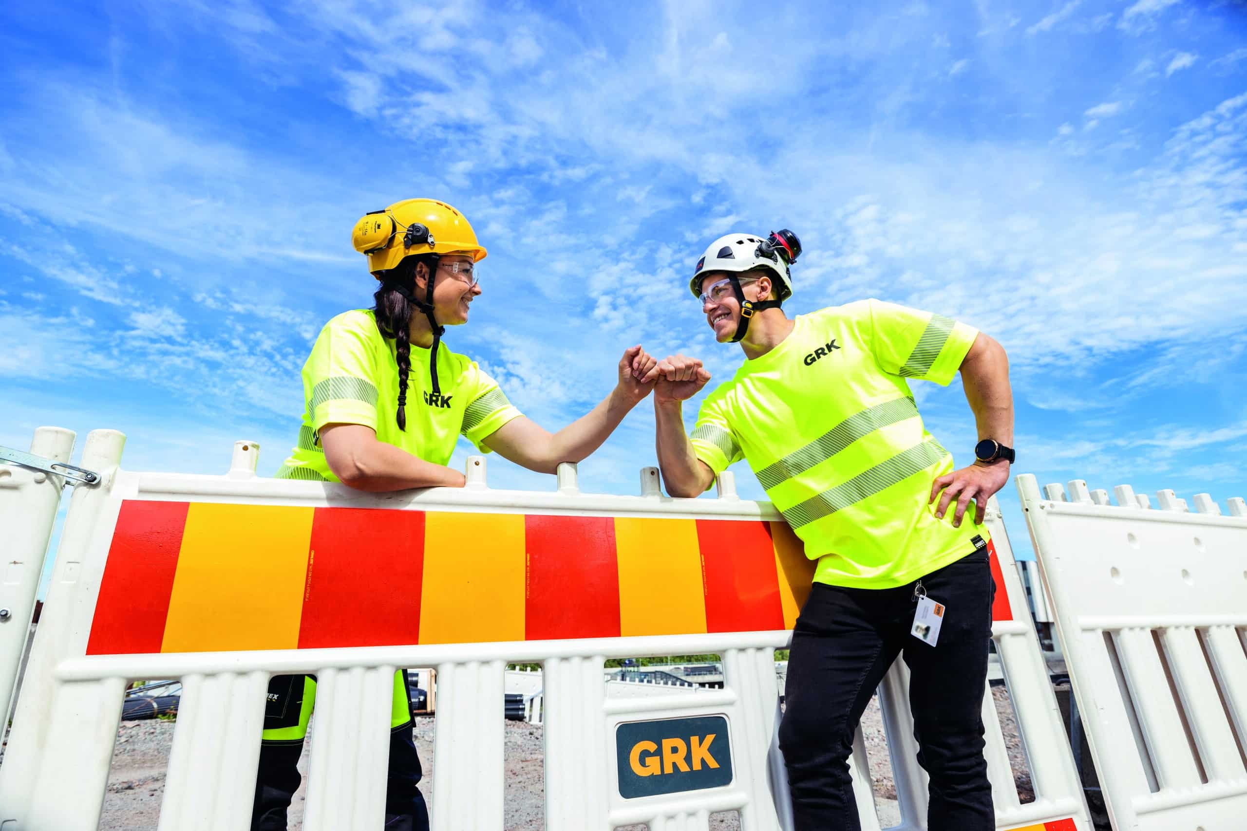 vå personer i neongula arbetskläder och hjälmar står vid en vägbarriär och gör en fistbump. Soligt väder och blå himmel i bakgrunden.