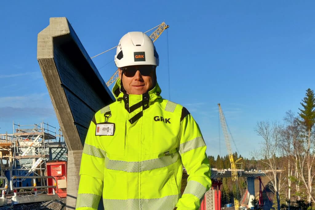 Person in neon yellow work clothes and white helmet stands in front of an ongoing bridge construction with cranes and forest in the background.