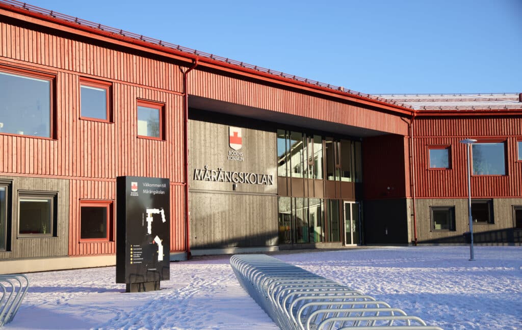 A modern school building in Boden with a red wooden façade and large glass windows. A sign at the entrance welcomes visitors to the school. 