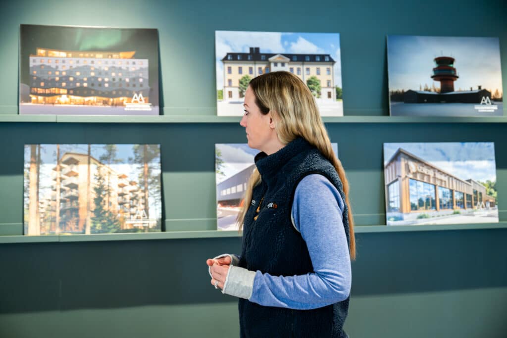 A woman in a blue fleece vest is standing in front of a green wall with shelves on which several architectural renderings of buildings are displayed. She looks at the pictures with her hands lightly clasped. 