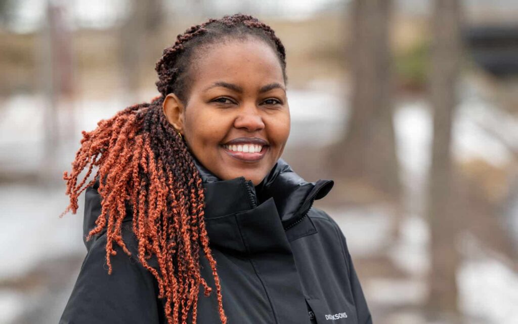 Christine Musembi står utomhus, ler stort och tittar in i kameran. Hon har långt, lockigt hår i en mörkbrun nyans med ljusa, kopparfärgade toppar, uppsatt i en hästsvans som faller över ena axeln. Hon är klädd i en svart, varm vinterjacka med hög krage och händerna ledigt i fickorna. Bakgrunden är suddig med en naturmiljö i milda, vinterlika färger.