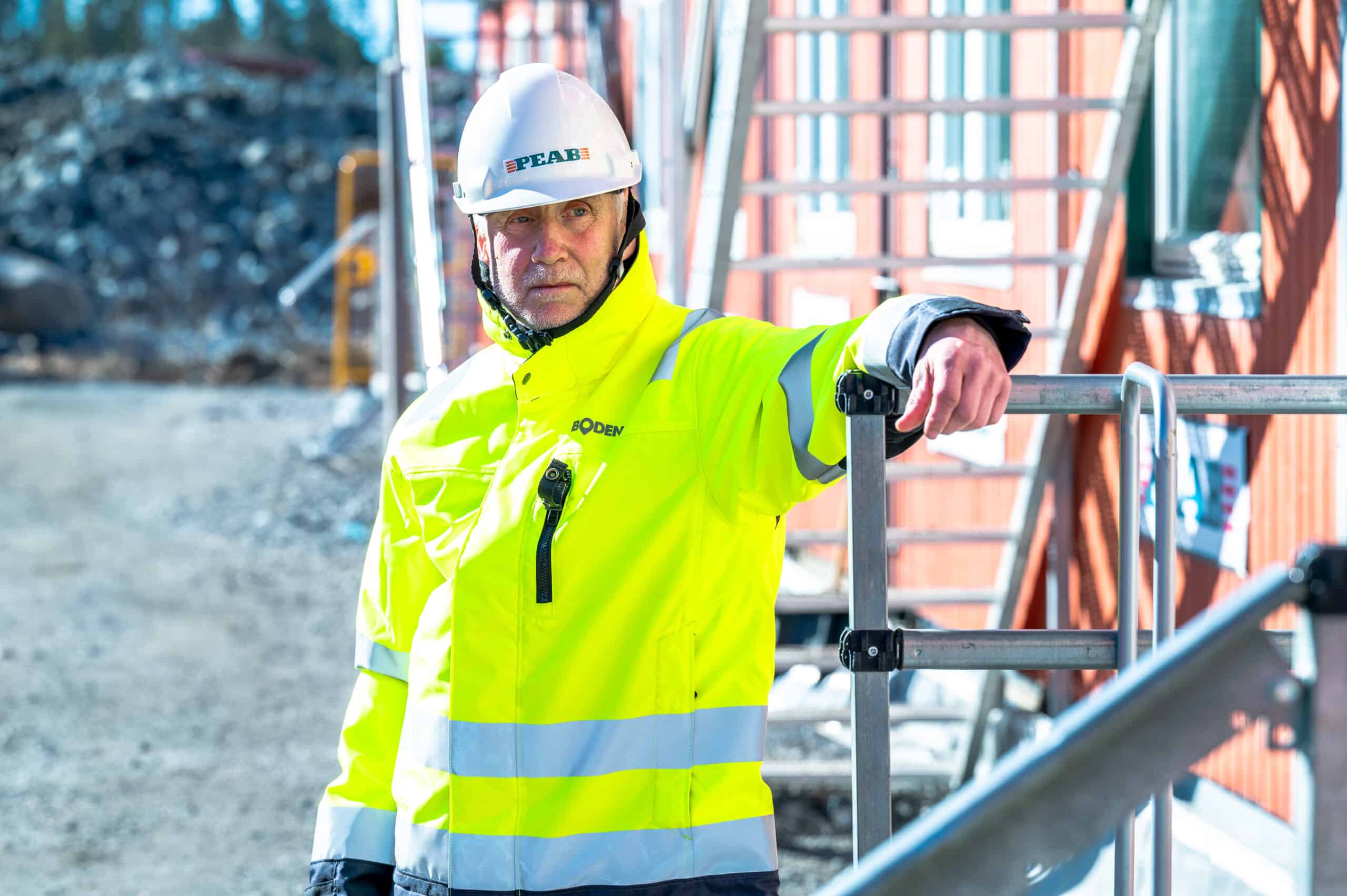 En byggarbetare i reflexjacka med texten “BODEN” och vit hjälm med logotypen “PEAB” står lutad mot ett räcke på en byggarbetsplats. I bakgrunden syns ett rött hus och byggnadsställningar.
