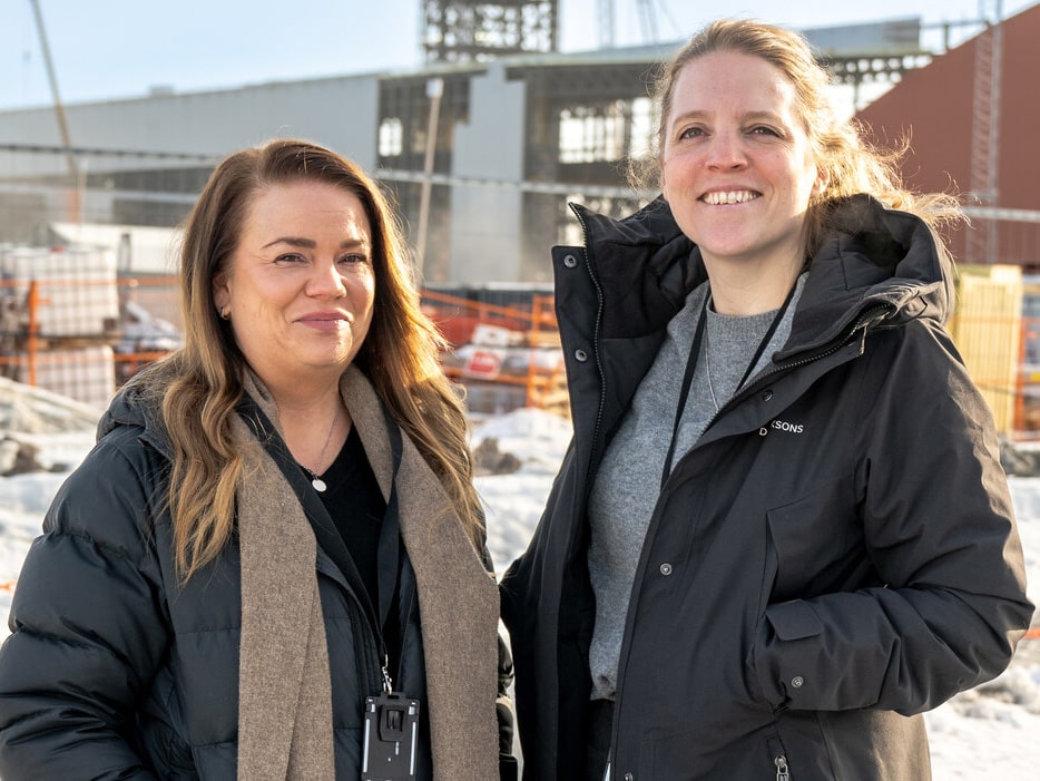 Två kvinnor står i vintermiljö framför ett stort industriområde under uppbyggnad. I bakgrunden syns byggkranar och delar av Stegras anläggning i Boden. Båda bär vinterjackor och ler mot kameran.