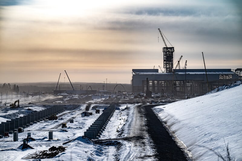 Vy över en stor byggarbetsplats i vinterlandskap. En lång väg leder in mot en stålbyggnad i uppförande, omgiven av kranar och byggmaterial. Himlen är dimmig i solnedgång eller soluppgång.