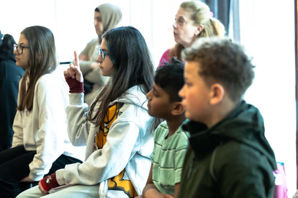 2 girls and 2 boys of different ages raise their hands to ask questions. In the background two female teachers, at the Stegra meeting place 