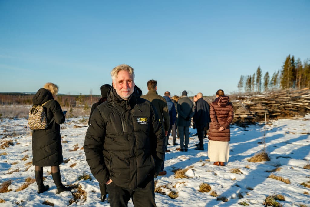 Mats Berg står leende i förgrunden med en grupp människor bakom sig på ett snötäckt kalhygge i Boden. Han bär en svart jacka med SM-veckan-logga. Den låga vintersolen skapar långa skuggor över marken.
