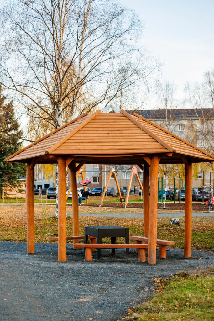  Ett öppet träpaviljongtak med sittplatser står i en höstlig parkmiljö. I bakgrunden syns en lekplats och bostadshus. En person gungar i ett rosa plagg.