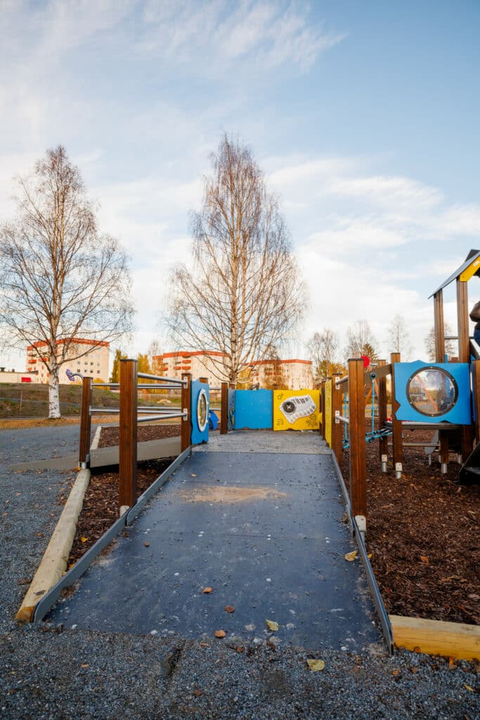 En tillgänglighetsanpassad lekplats med ramper och färgglada väggpaneler i blått och gult. I bakgrunden syns björkar och flerbostadshus. Bilden signalerar inkludering och barnvänlig stadsutveckling.