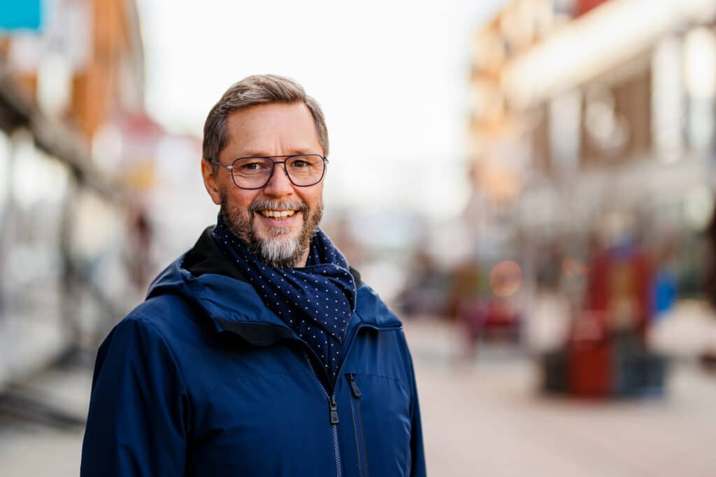 Magnus Åkerlund, kommunchef i Bodens kommun, står utomhus i stadsmiljö med oskarp bakgrund. Han bär glasögon, mörk jacka och en mörkblå scarf med vita prickar. Han ler mot kameran.