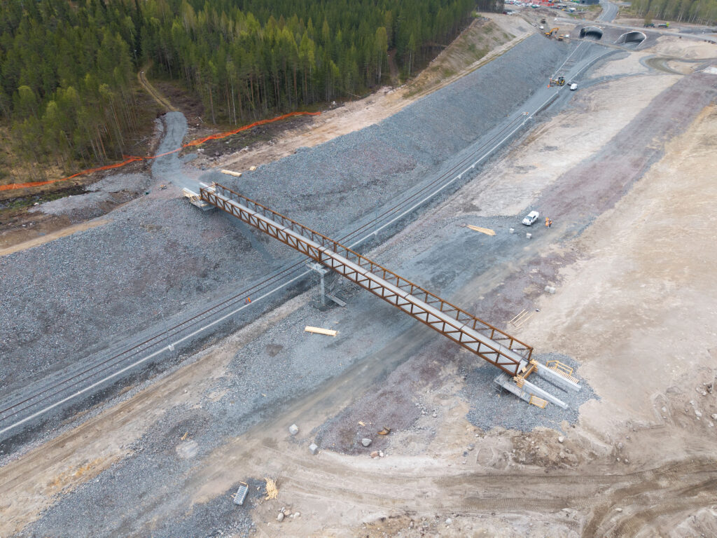 Drone image showing the pedestrian, bicycle and snowmobile bridge now in place over the railroad tracks at Boden Industrial Park. The bridge connects Södra Svartbyn with the Gruvberget recreational area, surrounded by forest, gravel and work vehicles. 