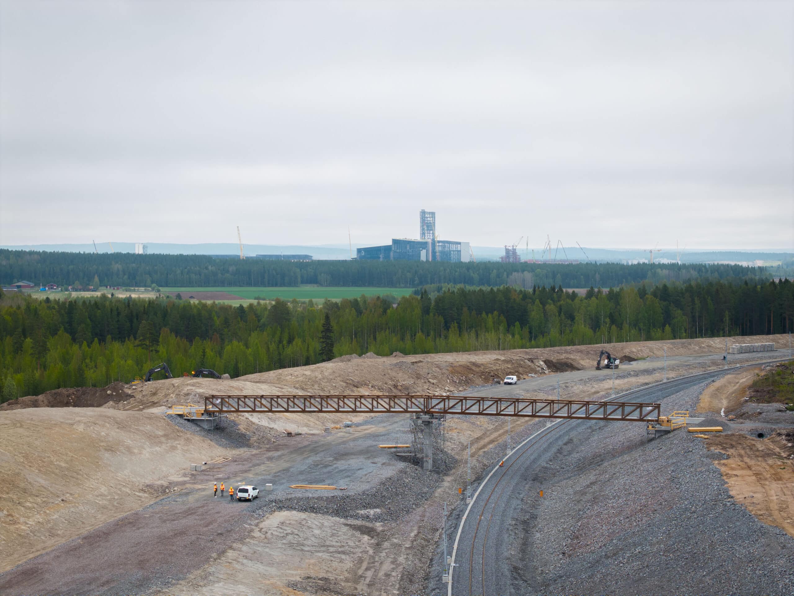 Vy över den nya bron som passerar över järnvägen med Bodens gröna landskap och Stegras anläggning i bakgrunden. Flera byggmaskiner och arbetslag syns i området.