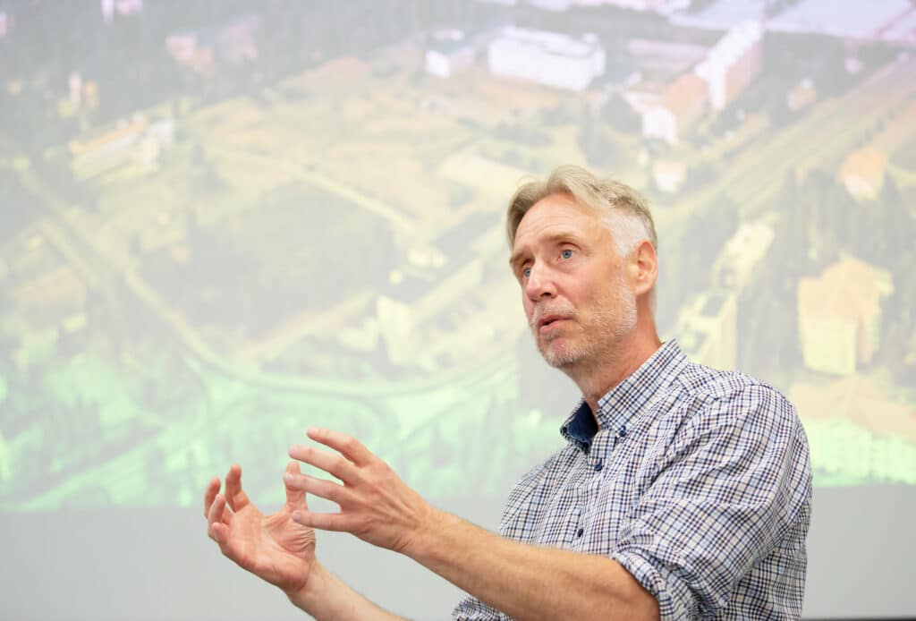 Mats Berg talar och gestikulerar engagerat framför en projicerad karta eller flygbild. Han bär en rutig skjorta och ser fokuserad ut. Bilden förmedlar strategiskt ledarskap i en planeringssituation.