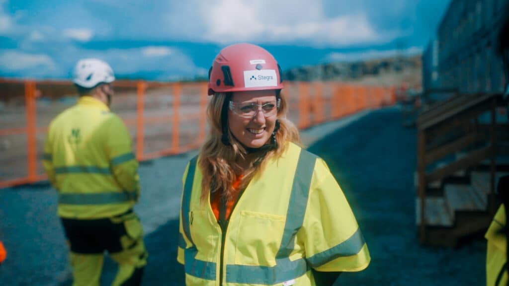Hedvig Paradis, ansvarig för vätgasteknik på Stegra, står utomhus i varselkläder och röd hjälm med företagets logotyp. Bakom syns byggarbetsplatsen i Boden.