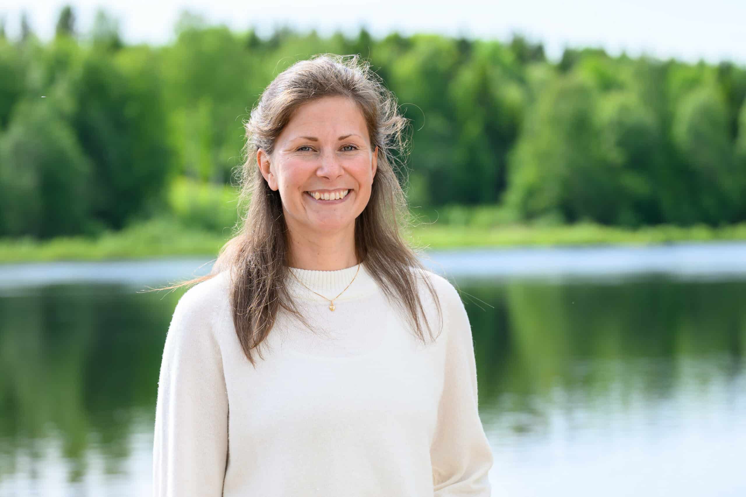 Lina Hällström står framför en sjö med skog i bakgrunden. Hon bär en ljus stickad tröja och halsband, och ler mot kameran i sommarljus.