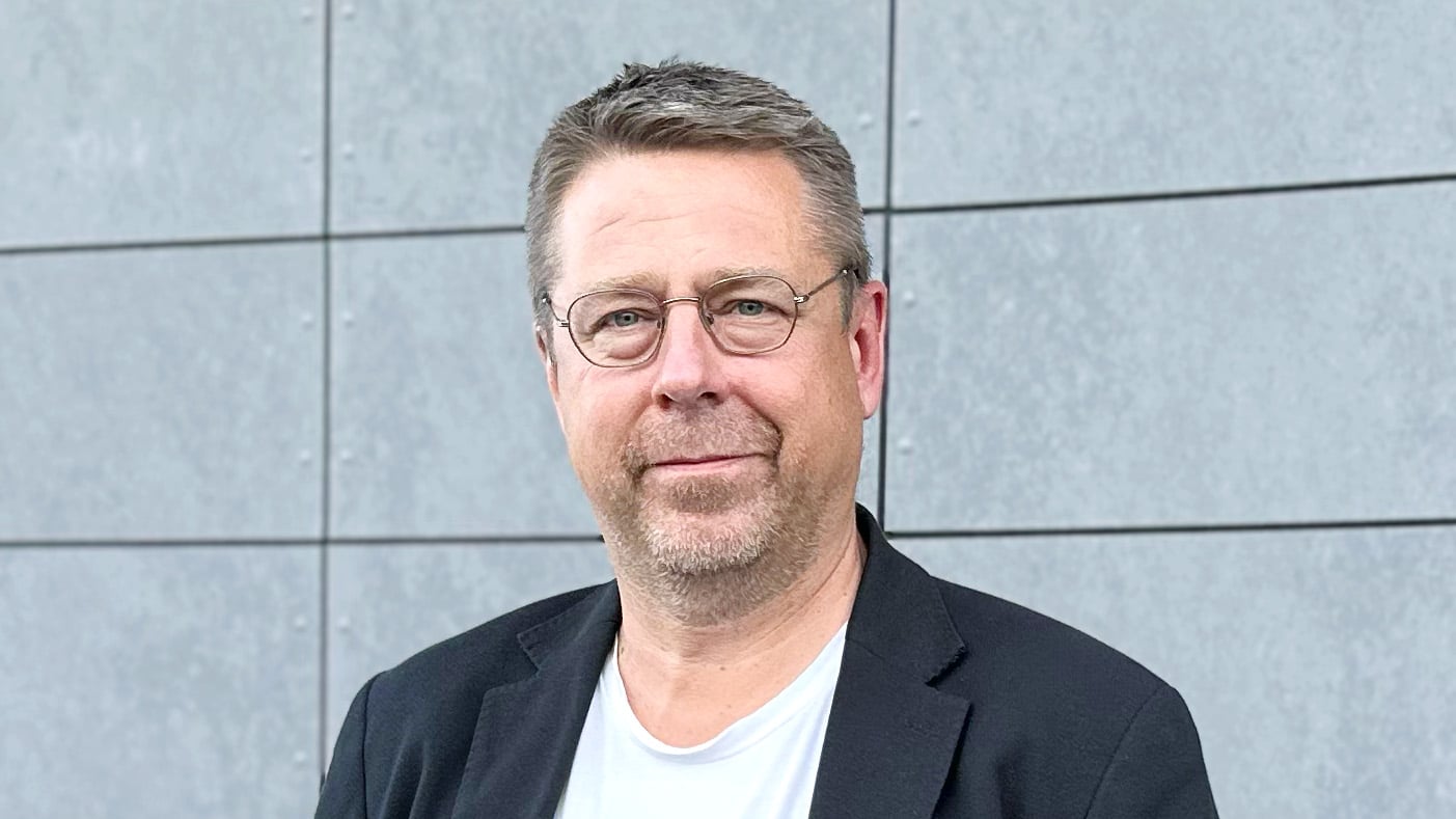 Thomas Sundqvist, ny utvecklingschef och biträdande kommunchef i Bodens kommun, fotograferad framför en grå fasad. Han bär glasögon, svart kavaj och vit t-shirt, och ser rakt in i kameran med ett lugnt uttryck.