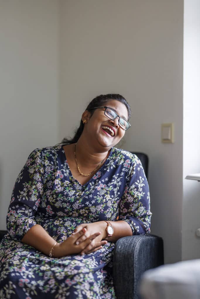 Naveena sits indoors in a chair and laughs, wearing a floral dress.