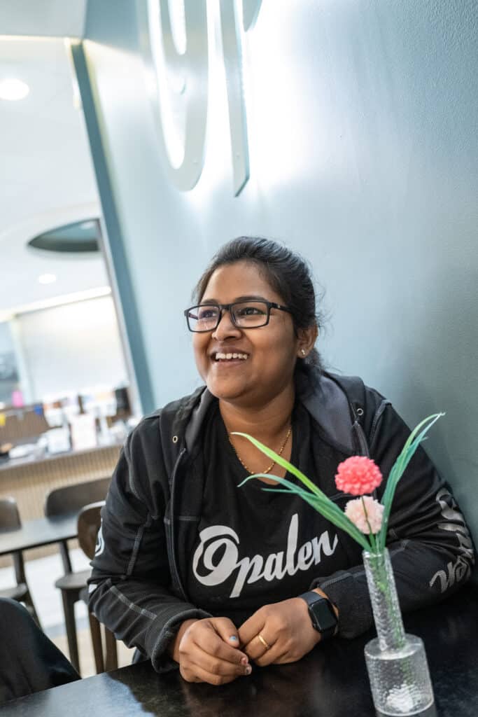 Naveena sits at a table, smiling, with a vase of pink flowers on the table