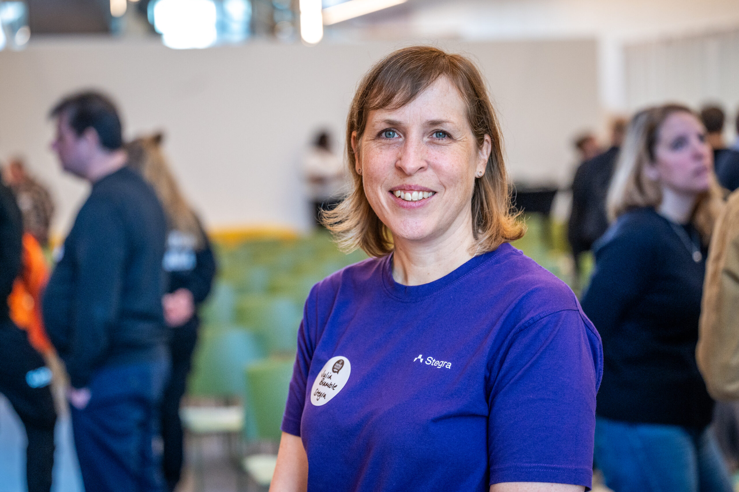 En kvinna i lila tröja tittar in i kameran och ser glad ut. På tröjan syns texten Stegra och en logotype.